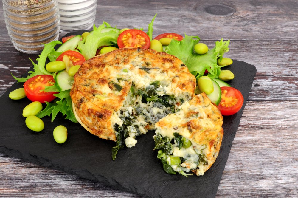 Quiche with spinach and cheese served on a black slate platter, garnished with cherry tomatoes and leafy greens, with salt and pepper shakers in the background.