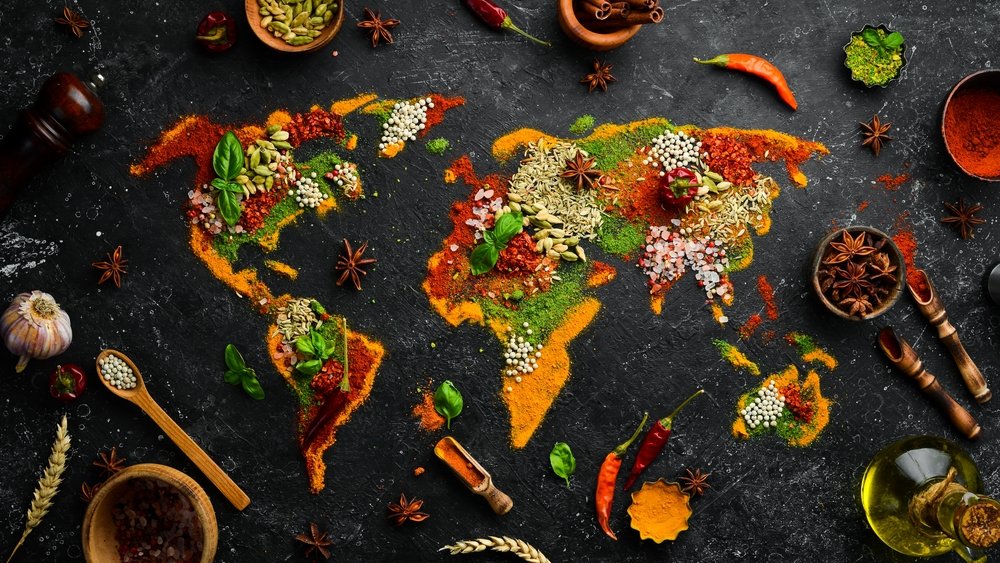 World map made of colorful spices and herbs on a black surface, with various spices, herbs, and cooking ingredients surrounding it.
